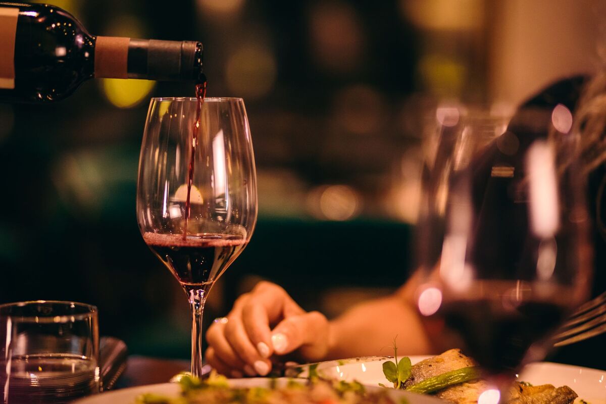 Close-up of red wine being poured into a glass at an elegant dinner setting, with a hand resting near a plate of gourmet food in a warmly lit restaurant.